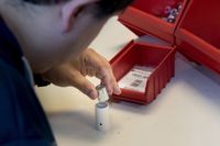 Iemand zit aan tafel en schuift 2 kleine onderdelen in elkaar. Op tafel staan 3 rode sorteerbakken met daarin zakjes en moertjes.