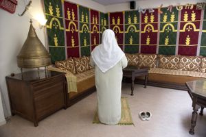 Vrouw in lang gewaad en hoofddoek om, staat op een kleed. Naast haar staan slippers. Ze staat in een ruimte met een hoekbank, wandkleden aan de muur en het woord 'welkom' aan de muur.