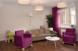 Huiskamer met houten vloer, een witte ronde salontafel, planten, een lichtbruine bank met kussens en twee leunstoelen met donkerroze bekleding en roze gordijnen