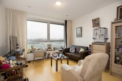 Appartement met grote ramen, een lichtroze leunstoel met gehaakt wit kleedje, een donkerbruine leren bank en kleine donkerhouten salontafel. Tegen de muur staat een vogelkooi.