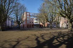 Vooraanzicht van een gebouw met rode bakstenen, vier verdiepingen aan een groot plein.