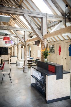 Een grote open ruimte in een oude boerderij met veel houten balken, met daarin een bar met glazen en tafels met stoelen.