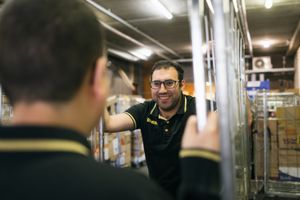 Twee mannen met zwarte polo's met geel Jumbo logo erop houden een metalen kar vast en kijken elkaar lachend aan. Ze staan in laad- en losruimte van een winkel tussen meerdere karren.