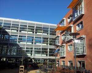 Gevel van een gebouw van 4 etages hoog. Recht voor is de gevel volledig van glas, haaks erop staan een gevel van rode baksteen en twee oranje zonneschermen boven de ramen