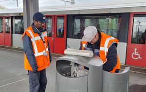 Twee mannen in oranje hesjes naast een metro op het perron en doen een nieuwe afvalzak in een prullenbak.