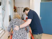 Je ziet van de achterkant een man in een donkerblauwe polo die een oudere dame ondersteunt op de loopbrug.