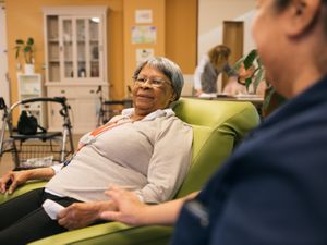 Over de schouder van een medewerker zie je een oudere vrouw liggen in een grote, lichtgroene stoel in een woonkamer. Ze kijkt in de camera.