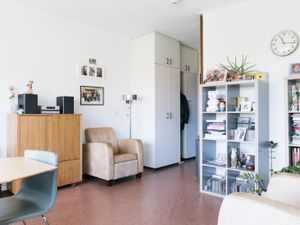 Een woonkamer met witte wanden, met links tegen de muur hoge witte kasten, een lage houten kast en een bruine stoel. Rechts tegen de muur hangt een klok en staat een open kast met vakken, met daarin beeldjes, boeken en cd's.