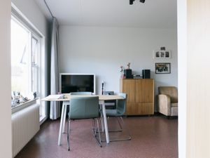 Een woonkamer met witte wanden, grijze gordijnen en een tafel met stoelen. Tegen de muur staan een televisie en daarnaast een houten kast.