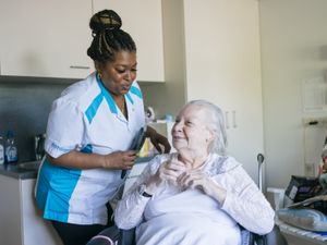 Een medewerker met blauw-wit werkuniform staat links naast een oudere dame in een rolstoel. De medewerker houdt een haarborstel vast en ze praten met elkaar.