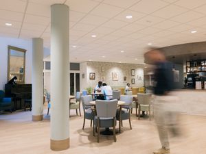 Een huiselijke ruimte met tafels, stoelen, een piano en oude schilderijtjes aan de muur.