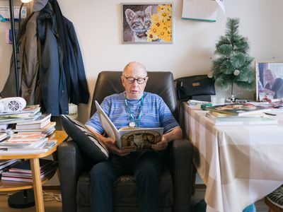 Een oudere man zit in grote, zwarte, leren stoel en leest een boek over katten. Aan beide kanten naast hem staan tafels met stapels boeken erop.