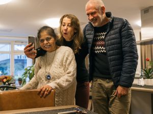 Drie mensen staan lachend naast elkaar in een woonkamer. Eentje maakt met een telefoon een selfie van hun drie.
