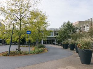Een oprit richting een gebouw met twee verdiepingen en witte deuren van een entree. Daarvoor bomen en grote bloembakken met bloeiende planten.