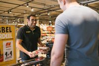 Een man met donker haar, een bril en een zwarte polo met de gele letters Jumbo erop staat met tomaten in zijn hand in een winkel. Hij kijkt lachend naar een andere man.