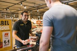 Een man met donker haar, een bril en een zwarte polo met de gele letters Jumbo erop staat met tomaten in zijn hand in een winkel. Hij kijkt lachend naar een andere man.