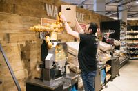 Een man met donker haar, een bril en een zwarte polo met een geel Jumbo logo erop staat in een supermarkt en vult een grote sinaasappelpers met sinaasappels.