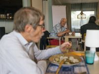 Bewoner zit een maaltijd te eten in de gezamenlijke woonruimte. Andere bewoner kijkt naar hem terwijl hij een toetje aan het eten is