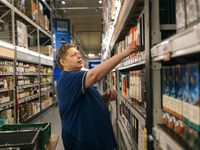 Een man met een blauwe polo staat in een groot gangpad met kasten vol drank. Hij haalt een fles drank uit een krat en zet deze in de stelling.