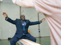 Een man in pak zit op een gym bank in een sportzaal. Hij doet de beweging na van de persoon die vóór hem staat.