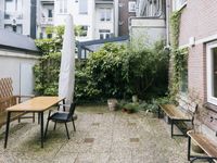 Stenen terras achter het huis met houten bankjes, een eettafel, stoel en een dichtgeklapte parasol