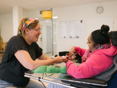 1 persoon in rolstoel doet lachend handoefeningen met een ander persoon die tegenover die zit