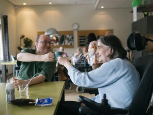 Een vrolijke man met bril proost met koffie met een oudere dame in scootmobiel. Ze zitten in een grote, gezellige ruimte met op de achtergrond ook mensen die koffie drinken.