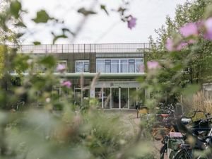 Door groene planten heen zie je een gebouw met twee verdiepingen en boven de ingang de tekst 'Hof van Sloten'. Daarvoor een rij geparkeerde fietsen.