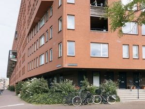 Een gebouw op de hoek van een straat, met rode bakstenen en 9 verdiepingen. Op de begane grond is buiten veel groen en staan fietsen geparkeerd.