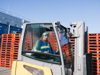 Een man met geel-blauwe jas en een pet zit buiten in een heftruck. Achter de heftruck staan grote stapels met rode kratten.