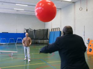 Een man in sportkleding en een man in een net pak gooien een grote rode bal over in een sportzaal.