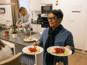 Een vrouw met kort, wart haar en spijkerblouse loopt vanuit de keuken de kamer in met twee borden fruit in haar hand.