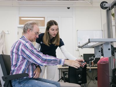 De fysiotherapeut begeleidt een man bij oefeningen met de handen. De man zit vóór een toestel met een beugel en gewichten.