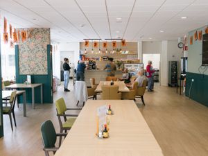 Een grote eetzaal met tafels en stoelen en een bar waar je eten en drinken kan bestellen. In de ruimte zitten en staan 6 mensen.