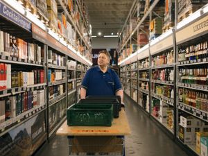 Een man met een blauwe polo met de gele tekst Makro erop loopt met een houten kar met kratten door een groot gangpad met kasten vol drank.