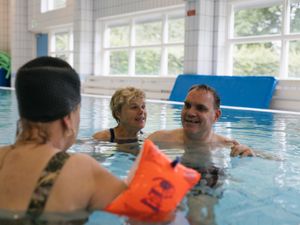Twee vrouwen en een man zwemmen in een zwembad omringd met grote ramen. Een van de vrouwen draagt zwembandjes.