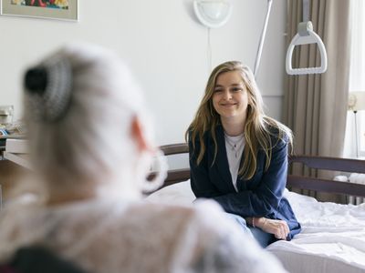 Een jonge vrouw met blond haar zit op een bed tegenover een oudere vrouw. Ze zijn in gesprek.