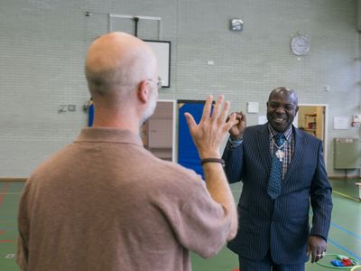 Twee mensen staan met hun gezicht naar elkaar toe in een sportzaal. De één draagt een polo shirt en de ander een blauw net pak.