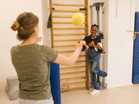 1 persoon oefent samen met een ander overgooien van een bal op 1 been staand