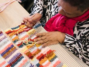 Een persoon met felroze sjaal werkt geconcentreerd aan een haakwerk met veel verschillende kleuren.