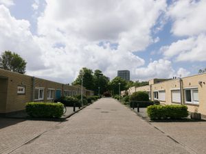 Een straat met aan weerszijden een gebouw van 1 verdieping. Op de achtergrond is een hoog gebouw en bomen te zien en een grote blauwe lucht met wolken.