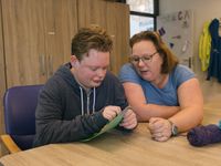 Twee mensen zitten aan tafel. De één borduurt en de ander begeleidt diegene hierbij.
