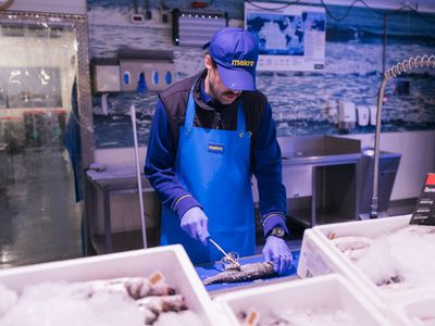 Een man met dikke jas, een blauw schort en blauwe pet met Makro erop staat in een kamer vol bakken met bevroren vis. Hij maakt een vis schoon.