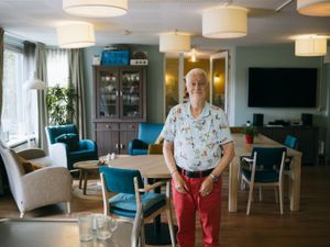 Een oudere man met rode broek staat in een gezellige woonkamer met veel lampen en blauwe stoelen en kijkt tevreden in de camera.