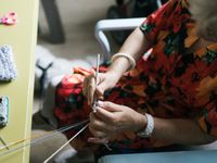 Je ziet van bovenaf een vrouw in een oranje bloemenjurk haken aan tafel.