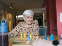 Een oudere dame zit in een grote kleurige ruimte en kijkt naar een spelbord Mens Erger je Niet op tafel.