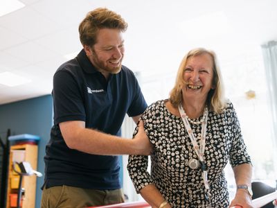 Een man in een donkerblauwe polo met Cordaan erop ondersteunt een oudere dame op de loopbrug. Ze lachen.