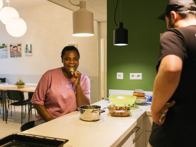 Een vrouw met roze shirt zit aan een kookeiland en eet lachend een stukje broccoli. In de keuken staat een lange, jonge man met een pet.