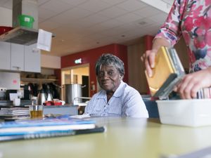 Een oudere dame zit aan tafel in een grote ruimte met een keuken. Voor haar staat een kop thee.