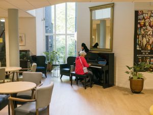 Een lichte, grote ruimte met tafels en stoelen en in het midden een vrouw achter een zwarte piano.